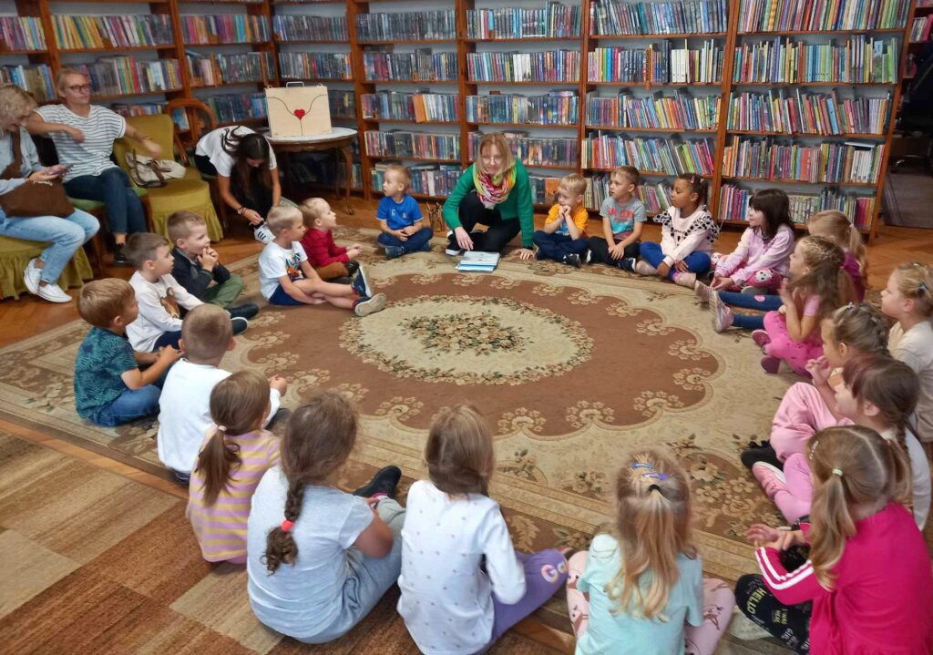 Duża grupa dzieci siedzi na dywanie. Między nimi siedzi bibliotekarka i coś mówi. Przed bibliotekarką leży książka.