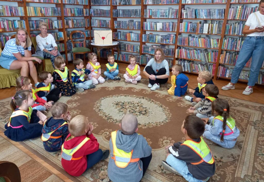 Duża grupa dzieci siedzi na dywanie. Między nimi siedzi bibliotekarka i coś mówi. 