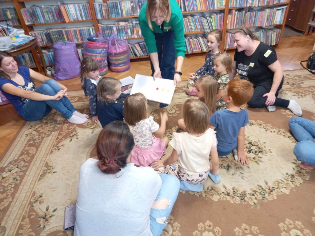 Dzieci i rodzice siedzą na dywanie i patrzą w stronę bibliotekarki, która. Bibliotekarka pokazuje ilustracje.