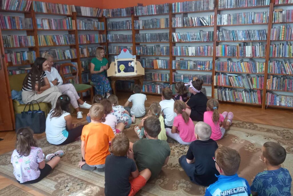 Sala biblioteczna. Dzieci siedzą na dywanie. Przed dziećmi na stoliku stoi drewniany teatrzyk. W otwartym teatrzyku znajduje się ilustracja. Obok teatrzyku siedzi bibliotekarka.