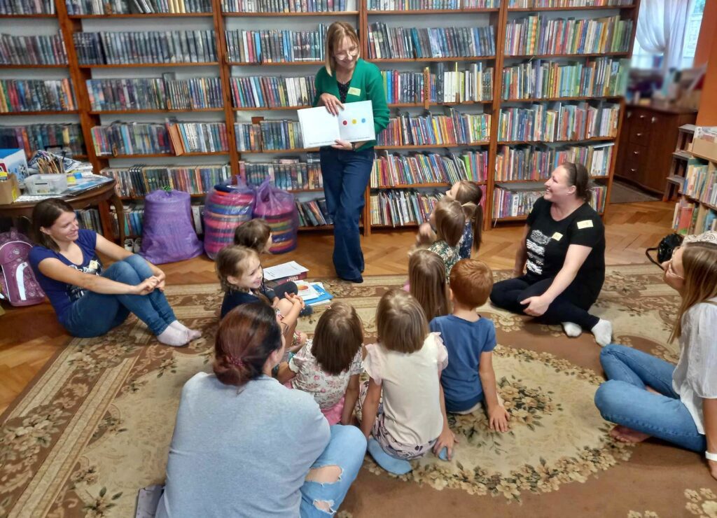 Dzieci i rodzice siedzą na dywanie i patrzą w stronę bibliotekarki, która. Bibliotekarka pokazuje ilustracje.