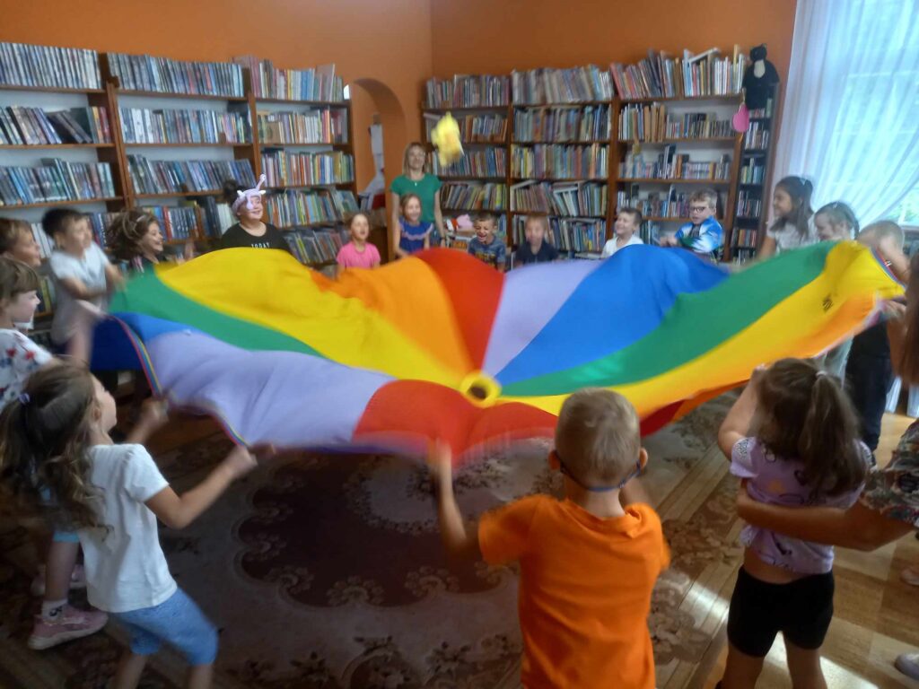 Sala biblioteczna. Dzieci, bibliotekarka i nauczyciele stoją w okręgu i poruszają kolorową chusta Klanzy.