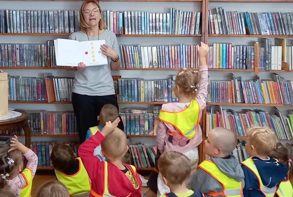 Grupa dzieci w sali bibliotecznej. Dzieci patrzą w stronę bibliotekarki pokazującej ilustrację w książce. Na ilustracji kolorowe kropki.