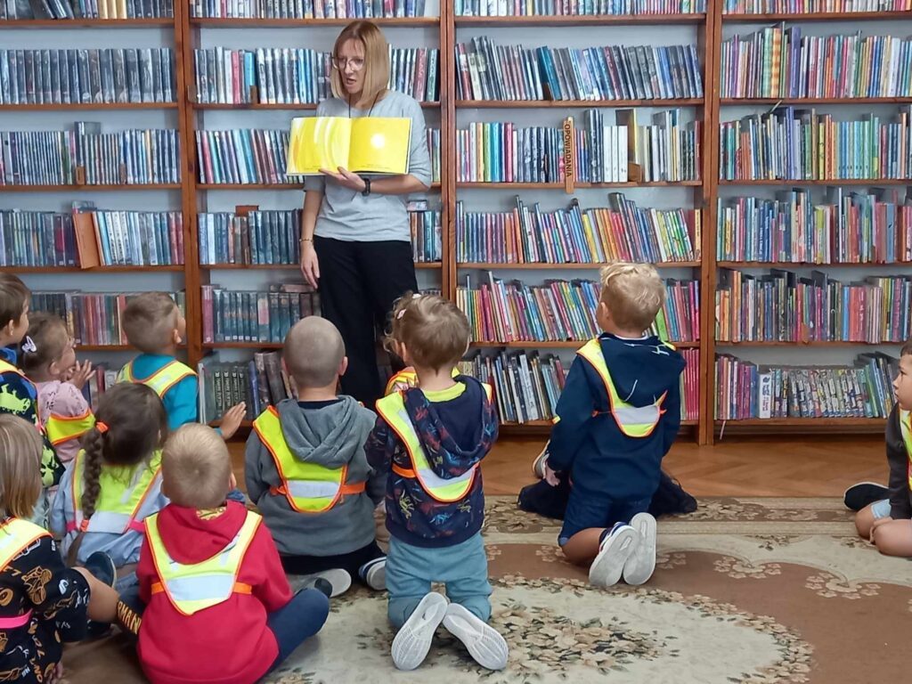 Grupa dzieci w sali bibliotecznej. Dzieci patrzą w stronę bibliotekarki pokazującej ilustrację w książce. 