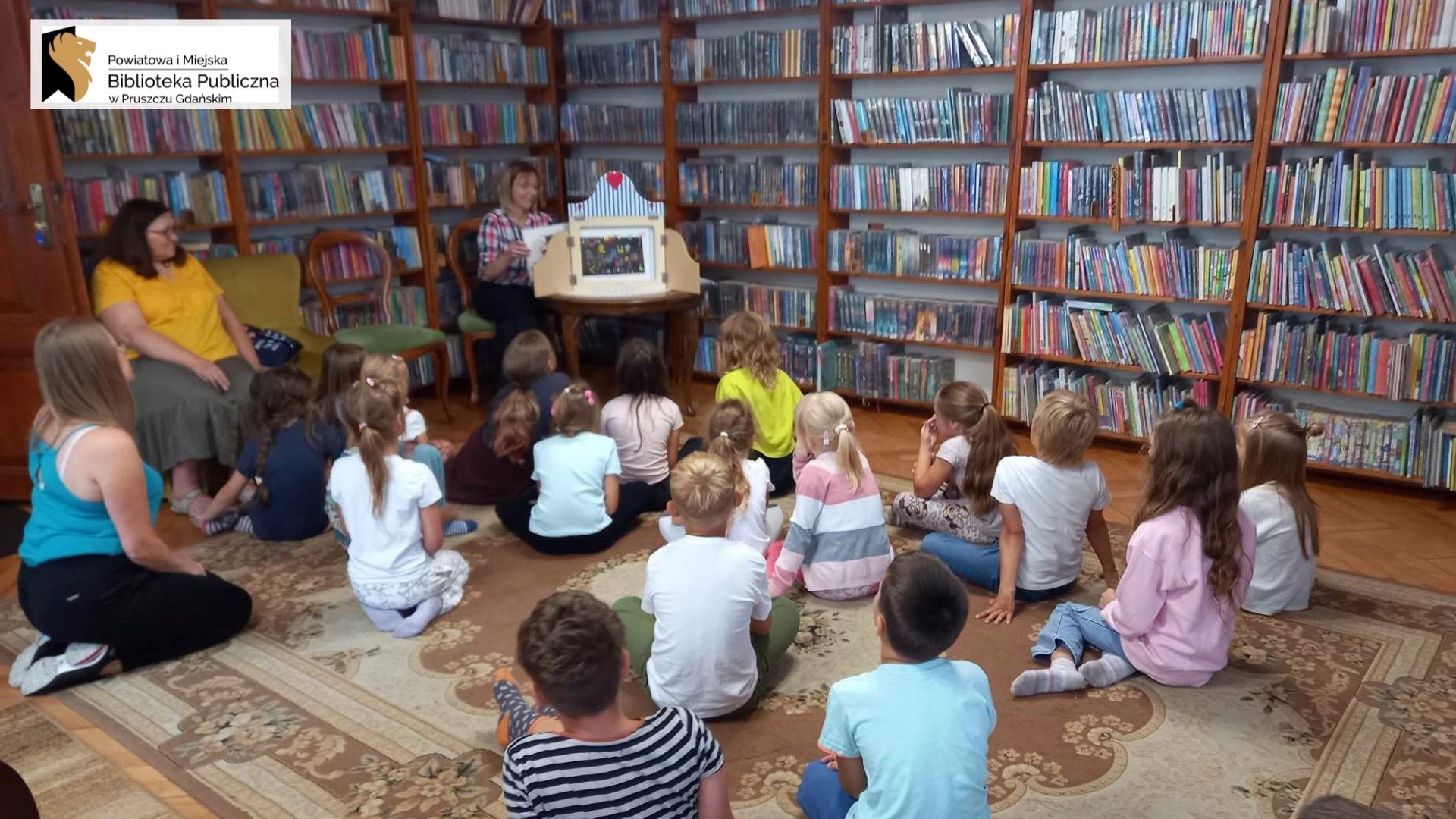 Sala biblioteczna. W tle regały z książkami. Dzieci siedzą tyłem na dywanie. Przed nimi bibliotekarka siedzi na krześle. Obok niej na stoliku skrzynka drewniana kamishibai.