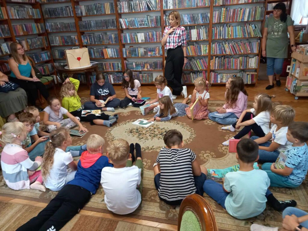 Sala biblioteczna. Na dywanie, w okręgu, siedzą dzieci. Dzieci trzymają w rękach książki. Między nimi stoi bibliotekarka i coś mówi.