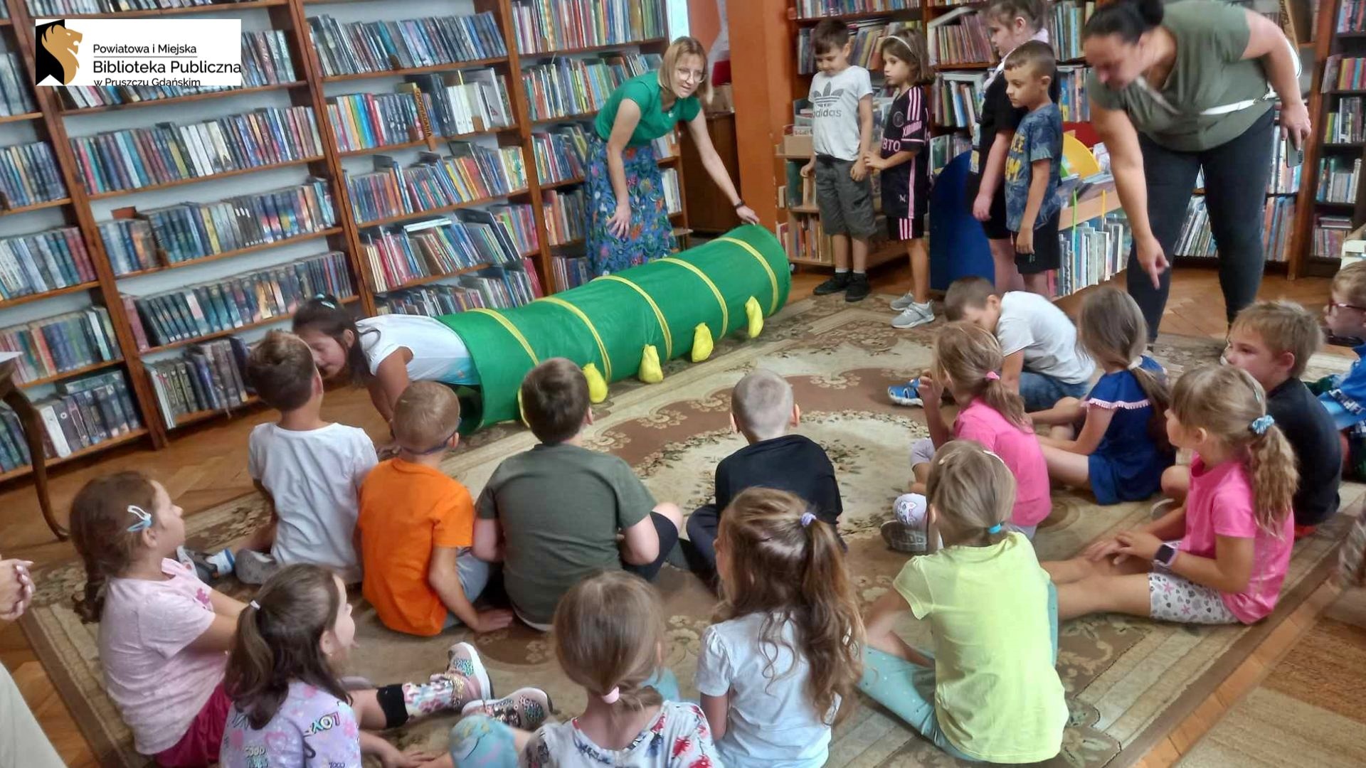 Grupa dzieci siedzi na dywanie w sali bibliotecznej. Kilkoro dzieci stoi w kolejce przez zielonym tunelem. Jedno z dzieci wychodzi z tunelu.