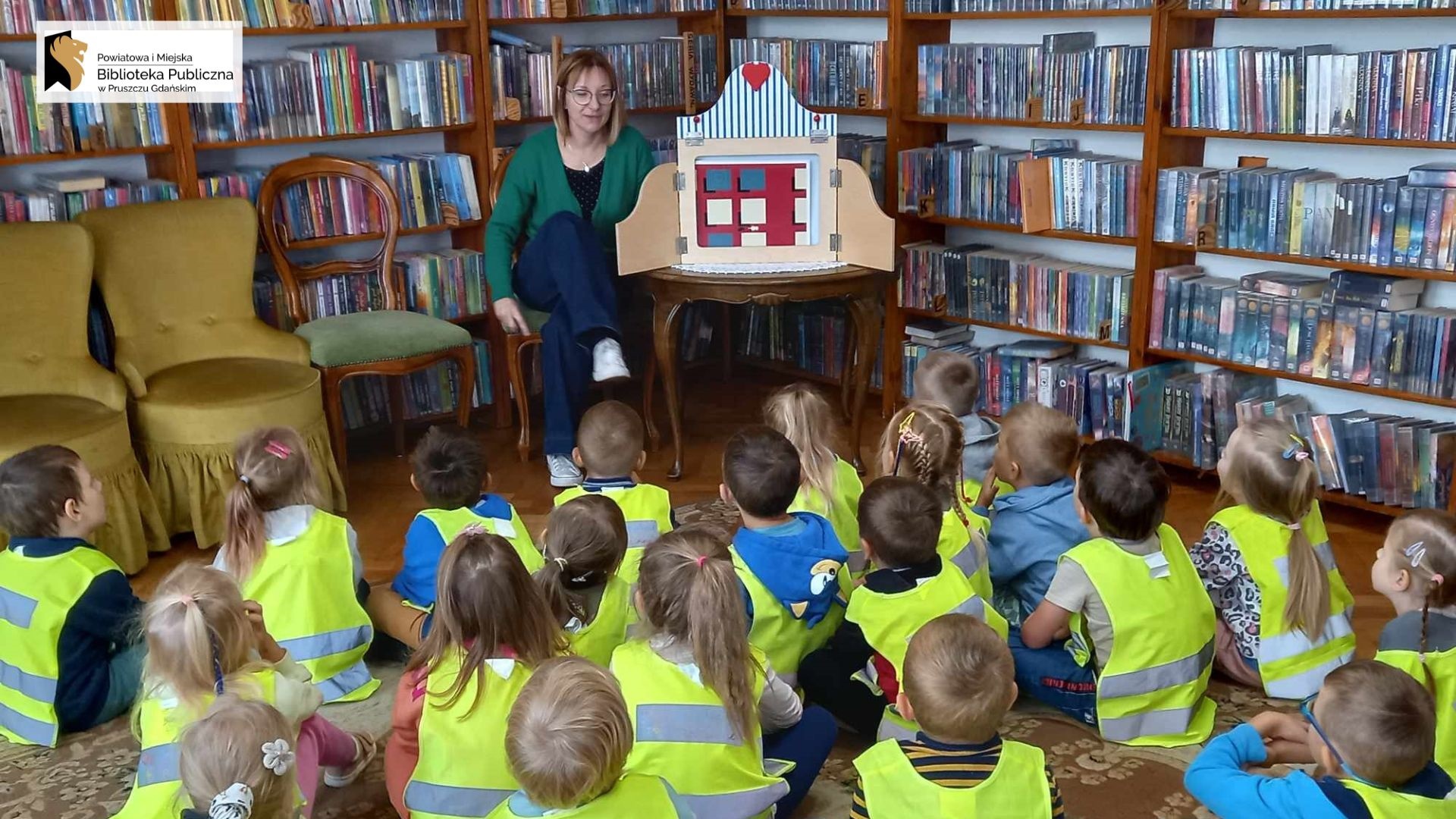 Duża grupa dzieci siedzi na dywanie. Dzieci patrzą w stronę siedzącej na krześle bibliotekarki, która coś mówi. Obok bibliotekarki stoi na stole otwarty teatrzyk. W teatrzyku ilustracja.