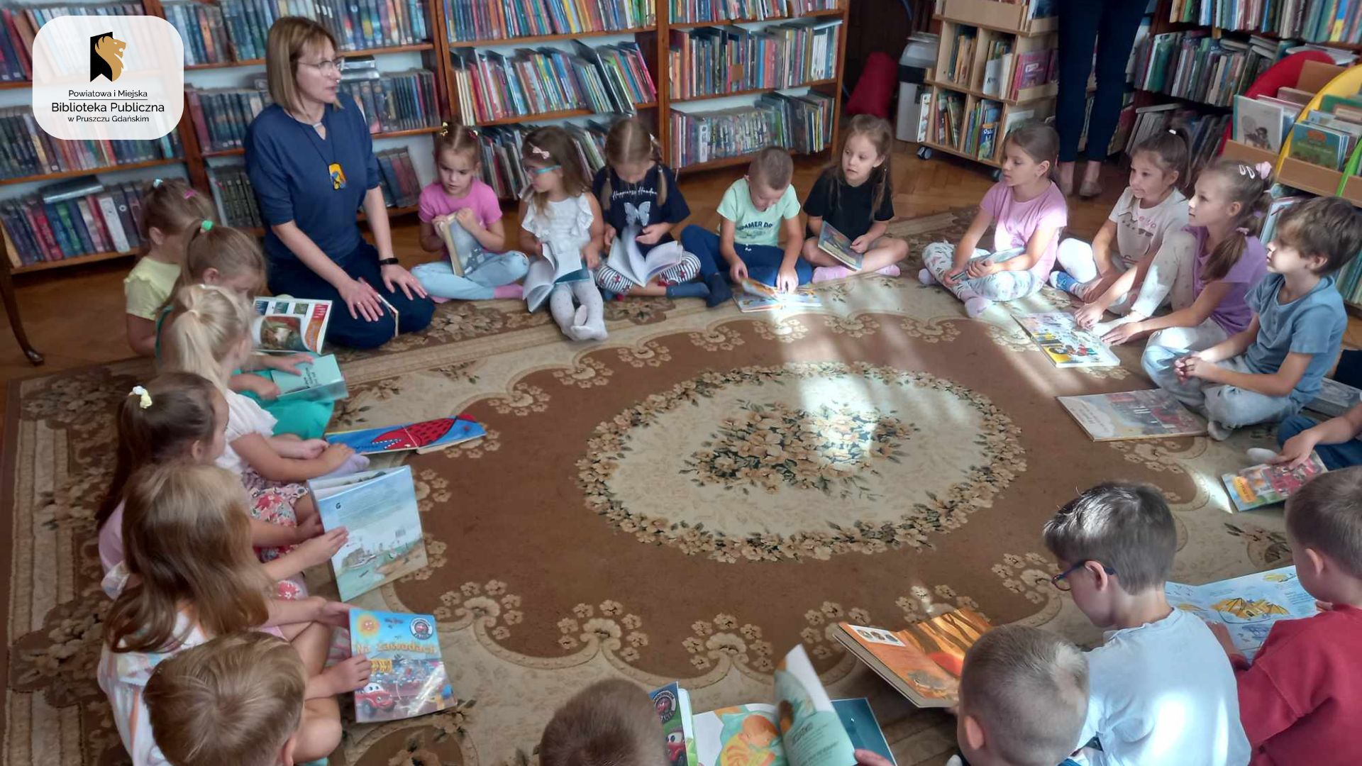 Grupa dzieci siedzi na dywanie. Między dziećmi siedzi bibliotekarka.