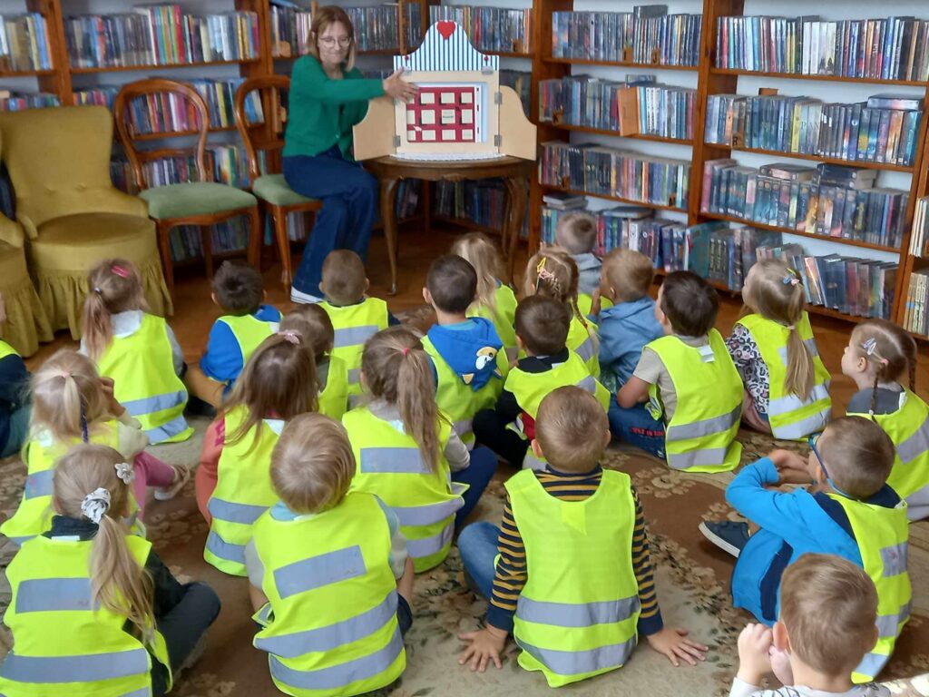 Dzieci siedzą na dywanie i patrzą na bibliotekarkę. Bibliotekarka siedzi na krześle. Obok niej na stole stoi drewniany teatrzyk kamishibai. W teatrzyku ilustracja.