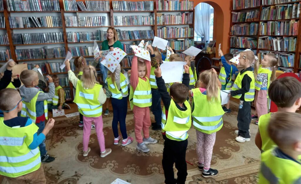 Grupa dzieci chodzi po sali bibliotecznej. Dzieci trzymają nad głową kartki z kolorowymi kropkami. Między dziećmi stoi bibliotekarka.