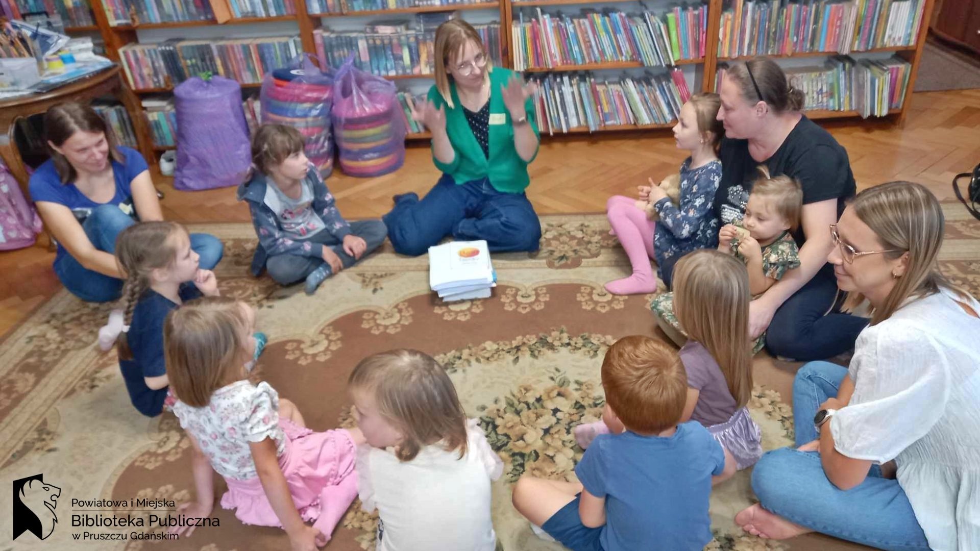 Na dywanie siedzi bibliotekarka, rodzice oraz małe dzieci. Bibliotekarka coś mówi.