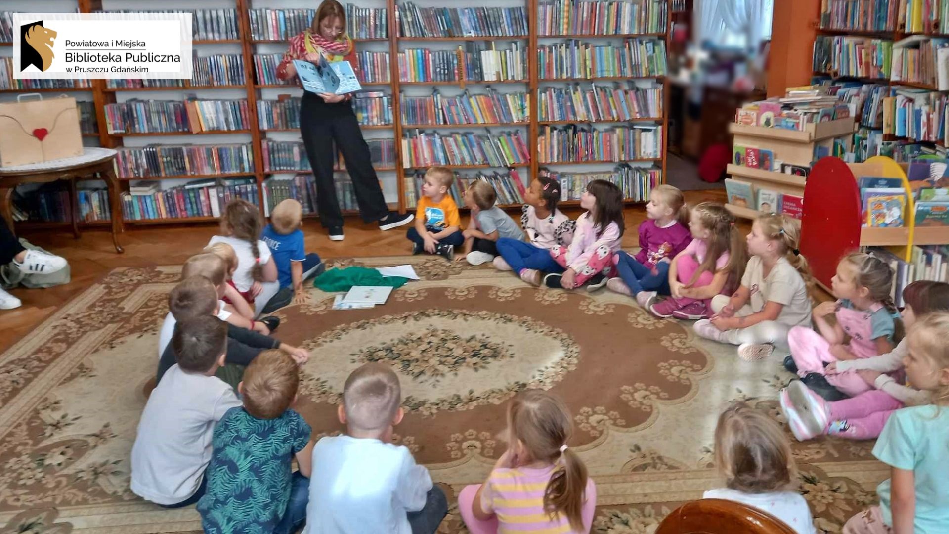 Grupa dzieci w sali bibliotecznej. Dzieci patrzą w stronę bibliotekarki pokazującej ilustrację w książce.