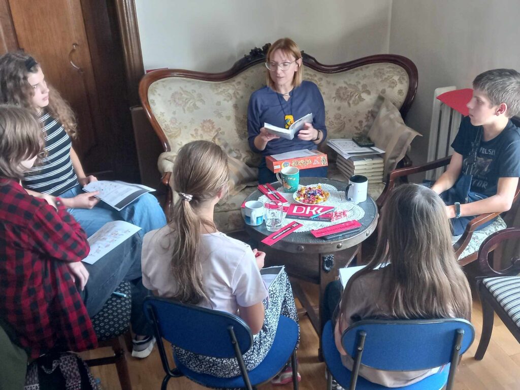 5 dzieci oraz bibliotekarka siedzą przy okrągłym stoliku. Na stoliku leża zakładki z napisem Dyskusyjny Klub Książki. Bibliotekarka trzyma w rękach otwartą książkę i coś mówi.
