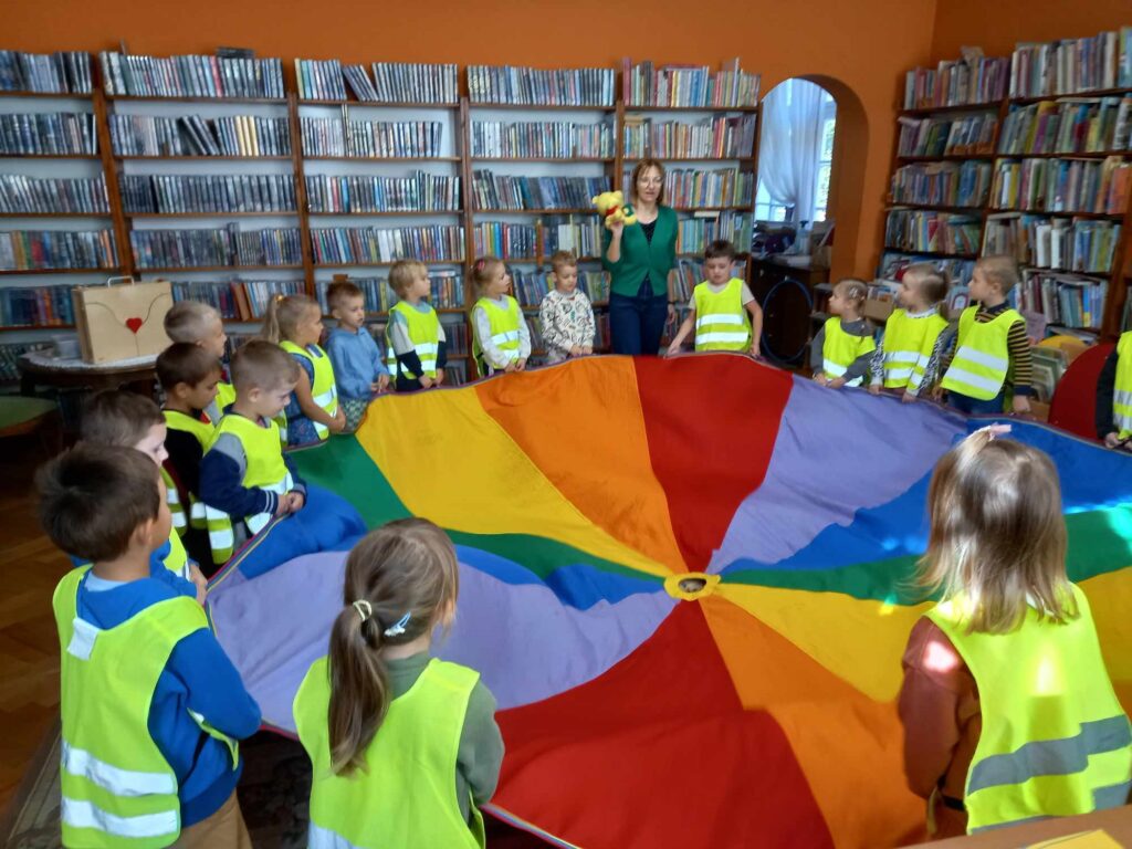 Dzieci stoją w okręgu i trzymają kolorową chustę. Między dziećmi stoi bibliotekarka z  misiem w rękach.