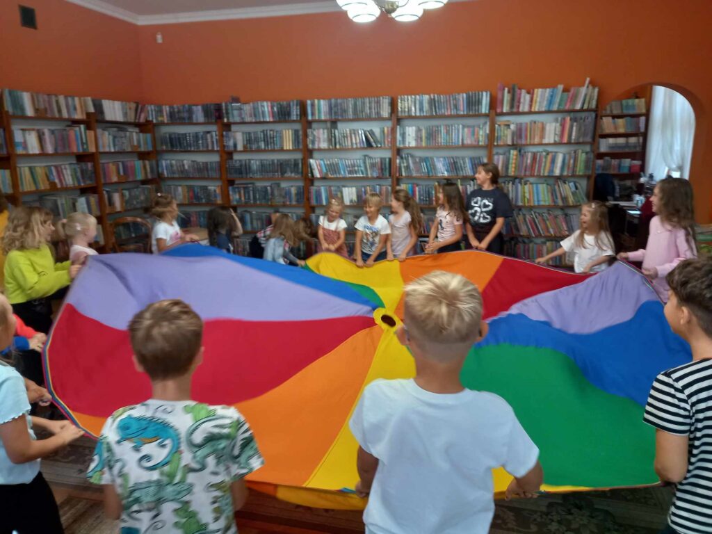 Sala biblioteczna. Dzieci stoją w okręgu i trzymają w rękach kolorową chustę.