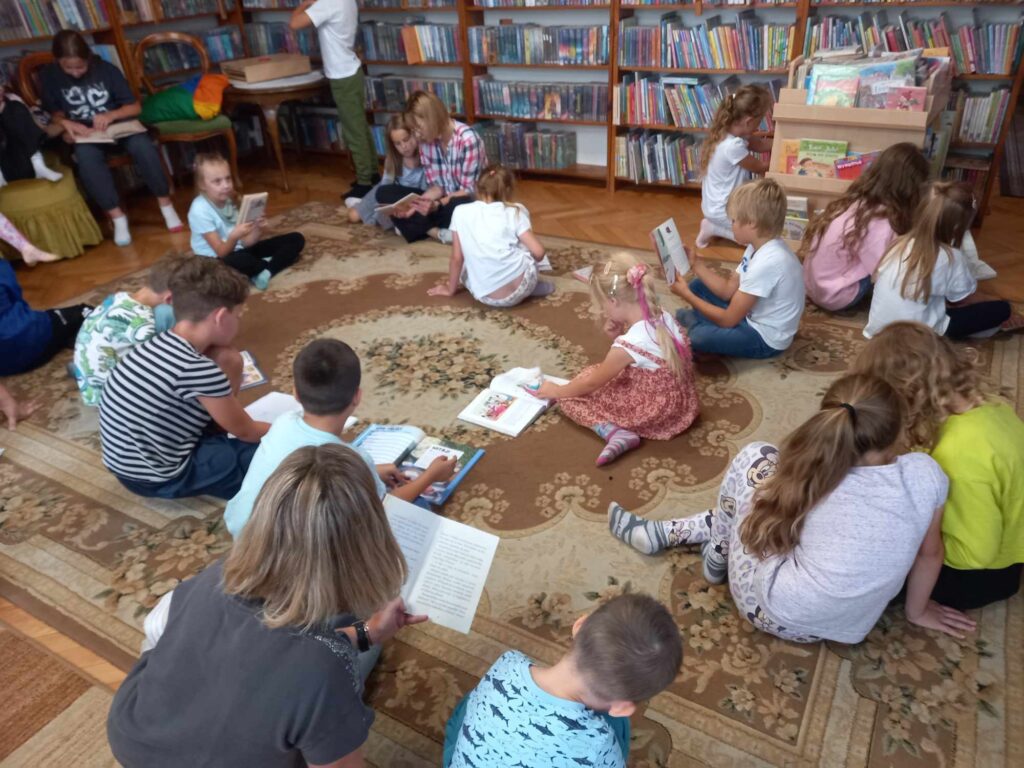 Sala biblioteczna. Na dywanie siedzą dzieci i oglądają książki. Między nimi siedzi bibliotekarka.