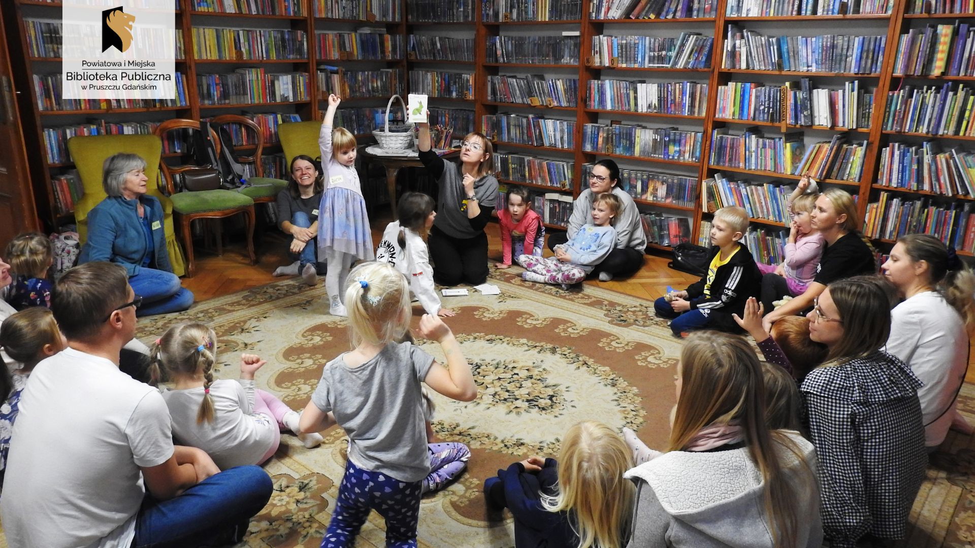 Duża grupa dzieci i dorosłych, w tym bibliotekarka, siedzą w kole. 2 dzieci stoi. Bibliotekarka trzyma w górze kartę przedstawiającą królika. 3 dzieci unosi rękę w górę. W tle regały z książkami.