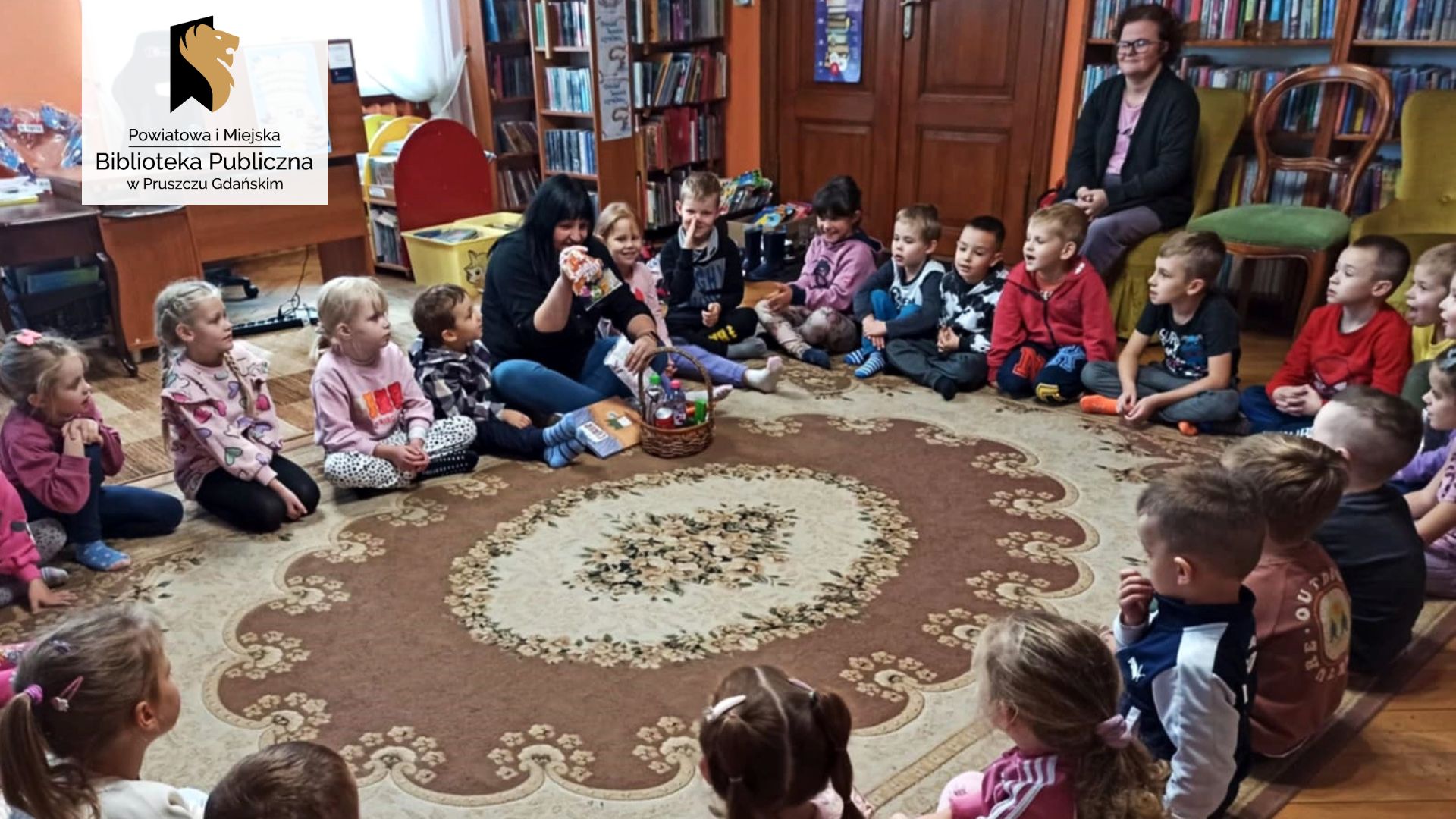 Duża grupa dzieci oraz bibliotekarka siedzą w kole na dywanie. Bibliotekarka trzyma w ręku opakowanie żelków. Przed nią leży kosz z różnymi produktami spożywczymi oraz książka pt. Sam cukier.