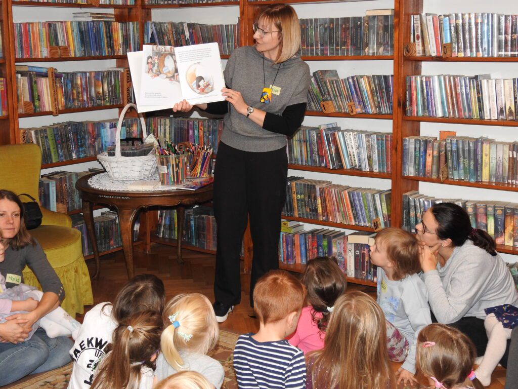 Bibliotekarka stoi, trzyma w ręku otwartą książkę. Przed bibliotekarką grupa dzieci i 2 dorosłych.
Za nią stolik z kredkami i koszem wypełnionym flamastrami oraz regały z książkami
