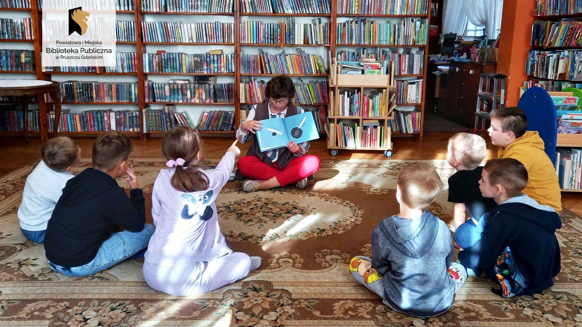 Bibliotekarka siedzi na dywanie i trzyma otwartą książkę. Naprzeciw niej siedzi 7 dzieci. Jedno z nich wskazuje palcem w kierunku książki. Na stronach książki nadrukowane są dwie czarne kropki i dwa białe prostokąty. W tle regały z książkami.