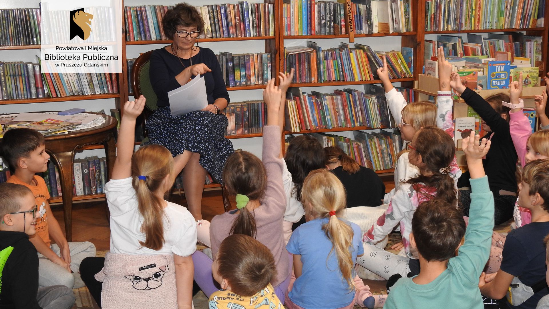 Duża grupa dzieci siedzi na dywanie. Są skierowani w stronę bibliotekarki siedzącej na krześle. Większość dzieci ma uniesioną rękę w górę. Bibliotekarka wskazuje palcem na jedno z dzieci. Za nią regały z książkami.