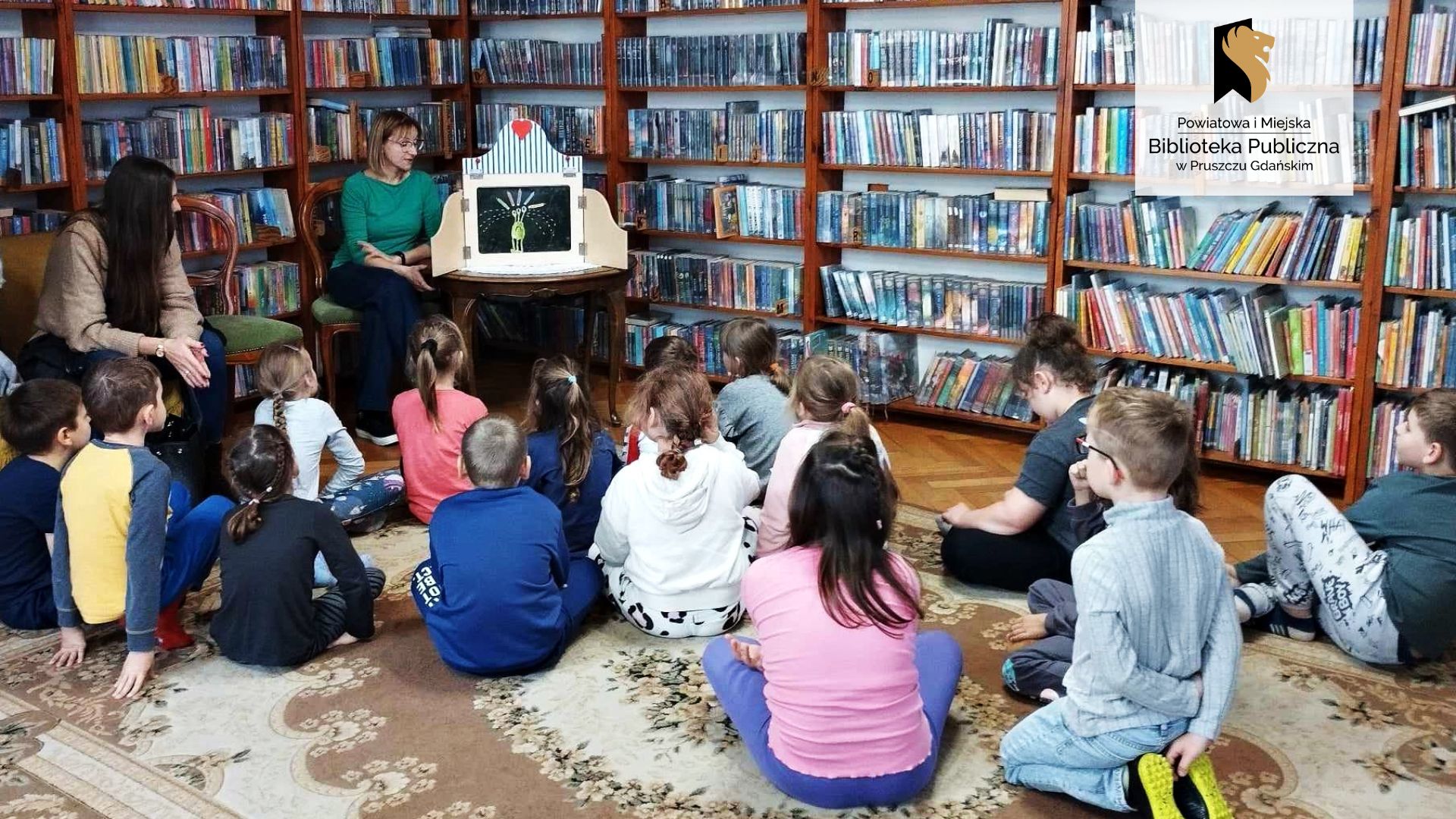 16 dzieci siedzi na dywanie. Obok na krześle siedzi osoba dorosła. Wszyscy są skierowani w stronę bibliotekarki siedzącej na krześle. Przy niej stolik, na którym stoi otwarta skrzynia teatrzyku kamishibai. Wewnątrz teatrzyku jest karta przedstawiająca zielonego kosmitę, który płacze. W tle regały z książkami.