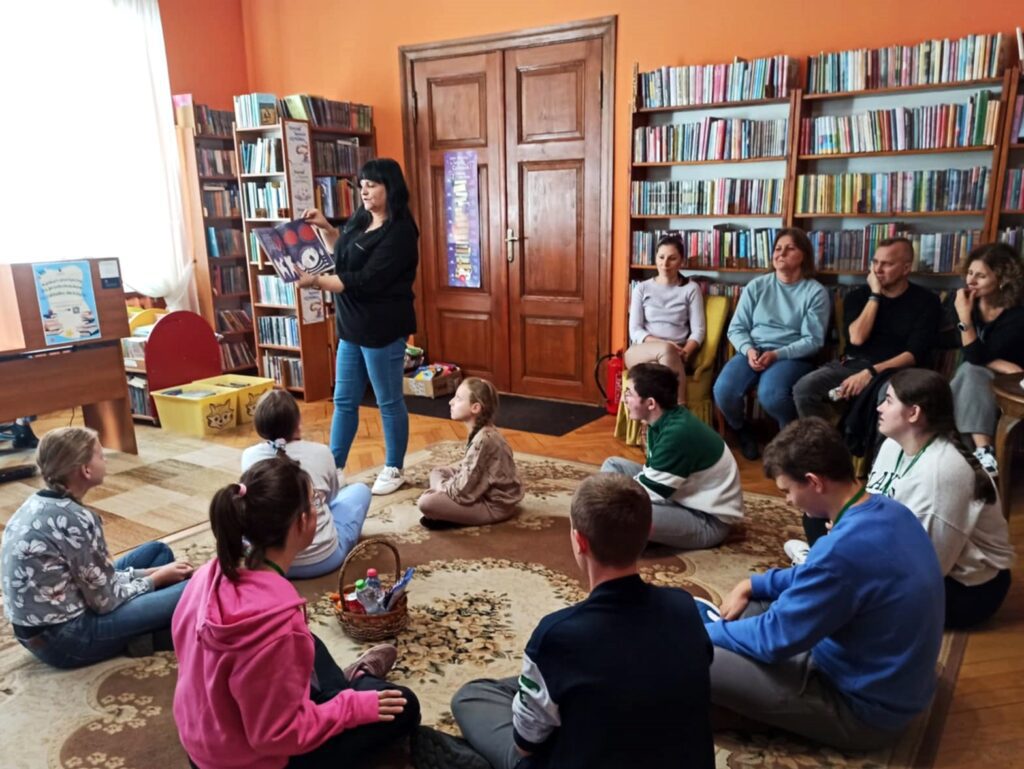 Bibliotekarka stoi. Trzyma w rękach otwartą książkę. W jej stronę zwrócona jest grupa 8 dzieci, które siedzą na dywanie. Obok dzieci, na krzesłach, siedzą 4 osoby dorosłe.