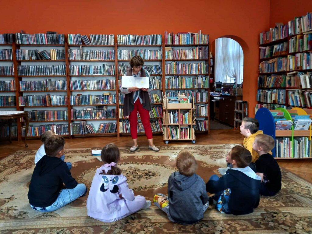 7 dzieci siedzi na dywanie i jest skierowanych w stronę stojącej bibliotekarki, która trzyma otwartą książkę. W tle regały z książkami.