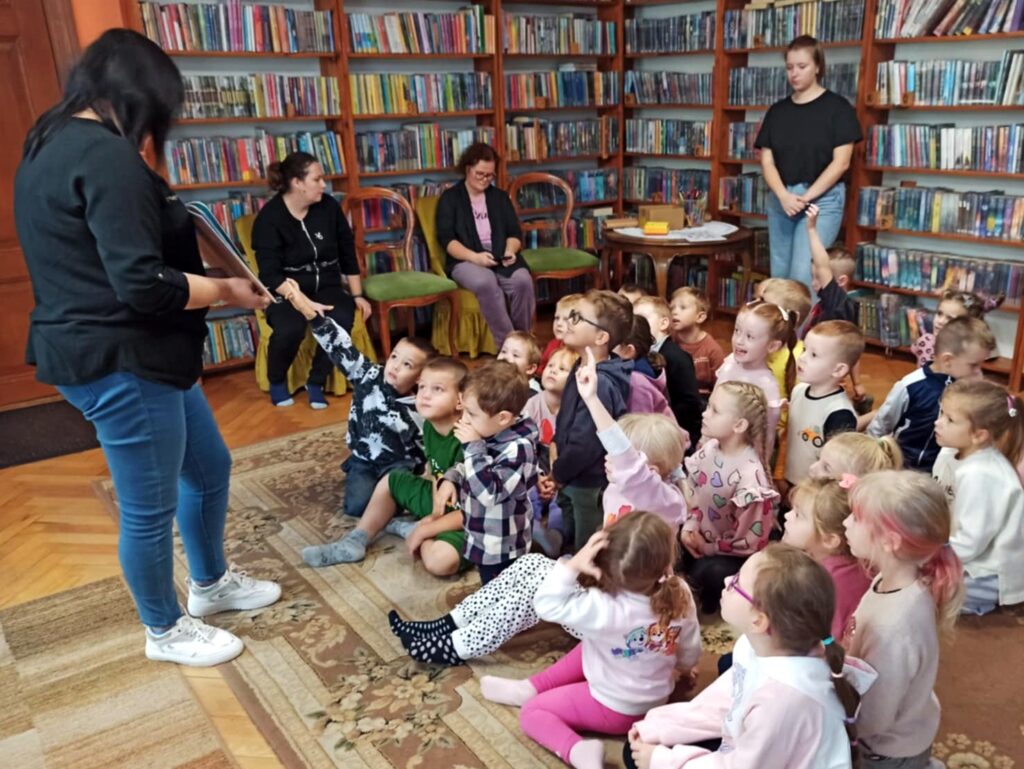 Grupa dzieci siedzi na dywanie. Jest skierowana w stronę stojącej bibliotekarki, która trzyma otwartą książkę. Troje dzieci ma uniesioną rękę w górze. Za dziećmi są trzy osoby dorosłe. Dwie siedzą na krzesłach a jedna stoi. 