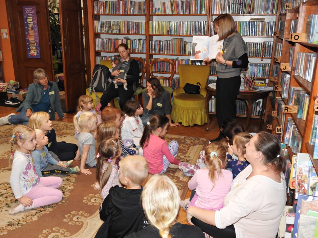 Duża grupa dzieci oraz kilkoro dorosłych są skierowani w stronę bibliotekarki czytającej książkę. W tle regały z książkami.