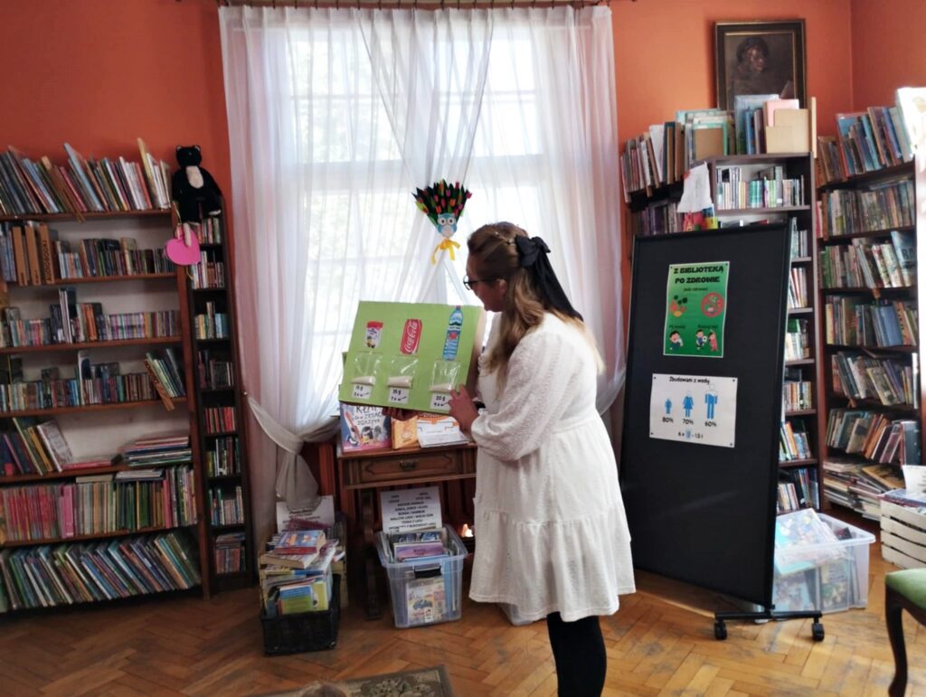 Bibliotekarka trzyma w rękach tablicę na której nadrukowane są 3 napoje tj. puszka coca coli i woda o smaku truskawkowym. Pod każdym napojem woreczek z cukrem. Za kobietą stand, na którym wiszą 2 plakaty. 