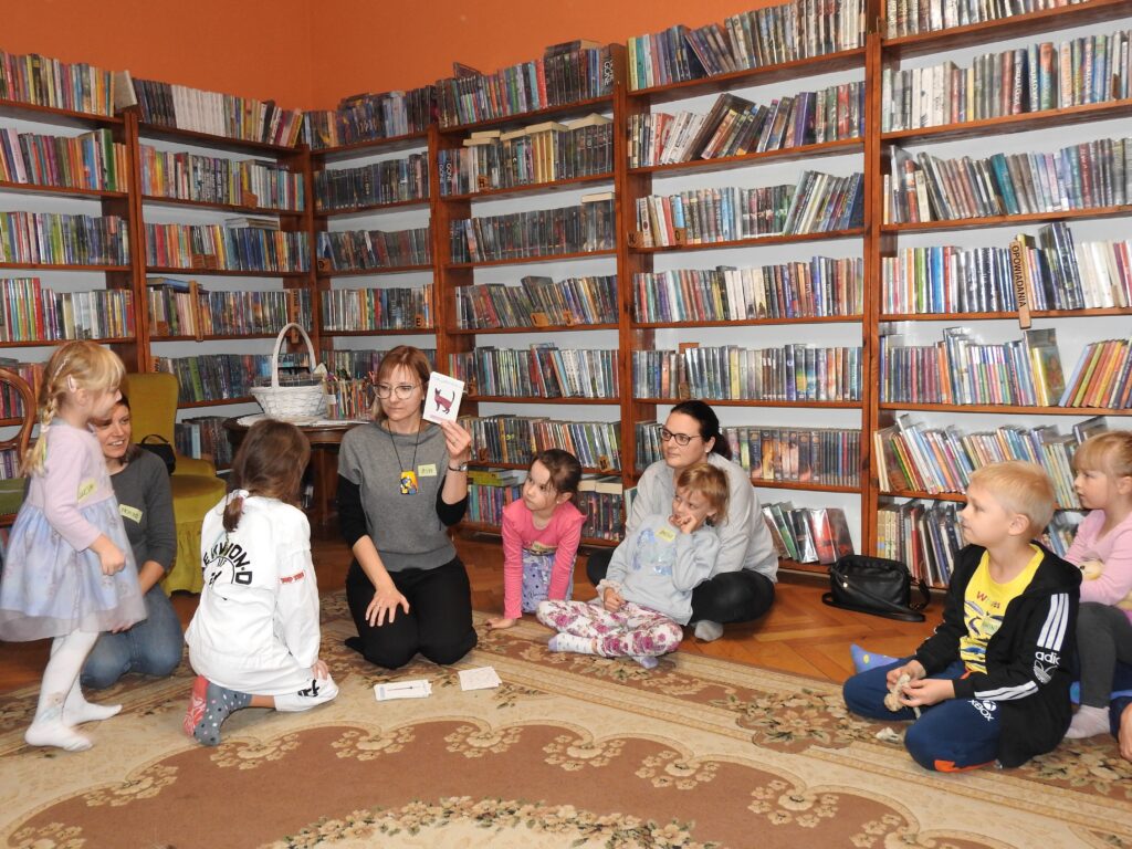 Bibliotekarka trzyma w dłoni kartę przedstawiającą kota. Obok niej 6 dzieci oraz 2 osoby dorosłe. W tle regały z książkami.