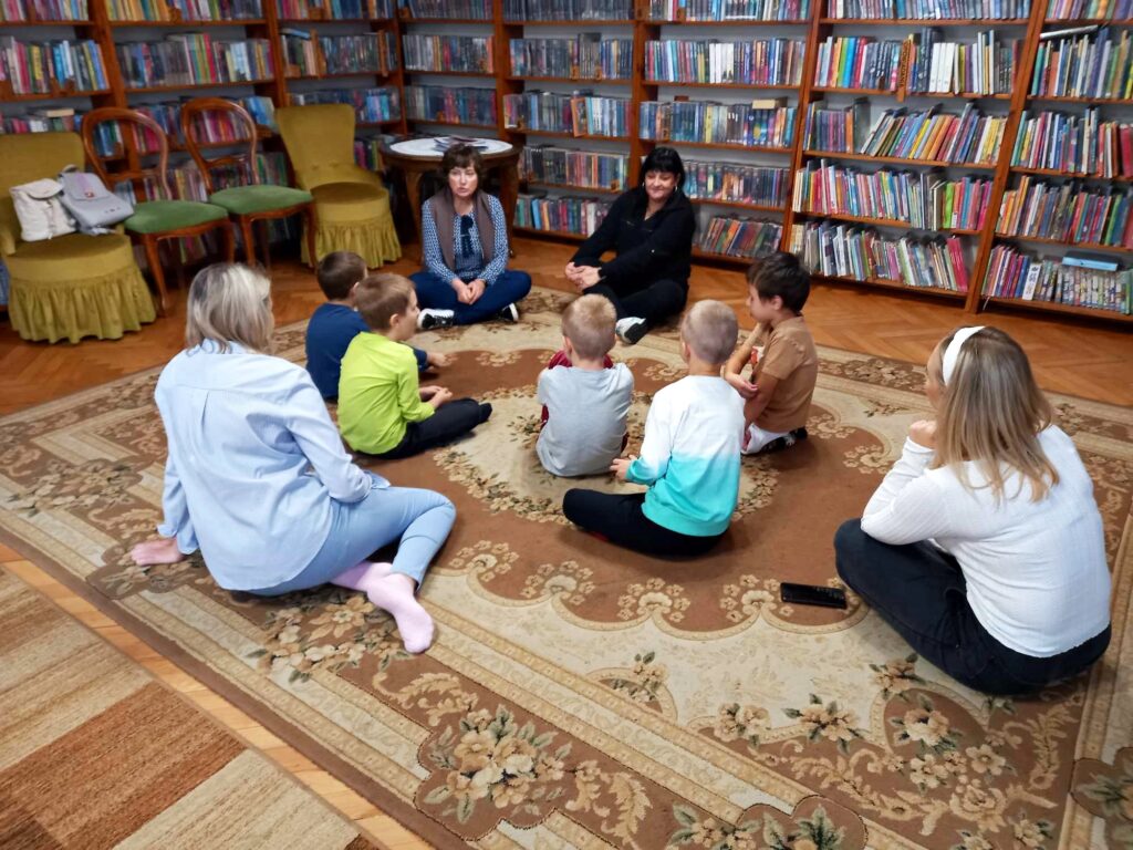 Dwie bibliotekarki siedzą na dywanie. Naprzeciw siedzi 5 dzieci oraz 2 osoby dorosłe. Z tyłu 4 krzesła, stolik oraz regały z książkami.