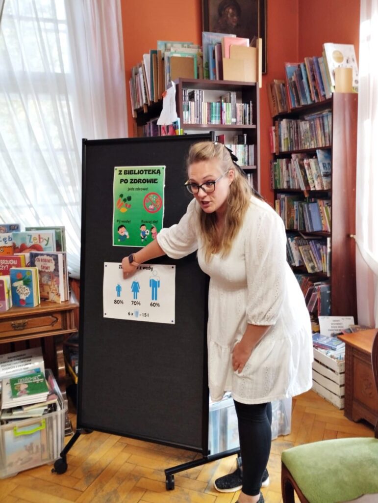 Bibliotekarka stoi przy standzie, na którym wiszą dwa plakaty. Na pierwszym napis: z biblioteką po zdrowie oraz grafiki warzyw i owoców, przekreślonych słodyczy, dziecka pijącego wodę, trzymającego piłkę oraz rozciągającego się. Na drugim plakacie napis: zbudowani z wody. Poniżej grafiki trzech postaci różnej wielkości, których wnętrze jest wypełnione na niebiesko. Bibliotekarka wskazuje palcem na jedną z grafik.