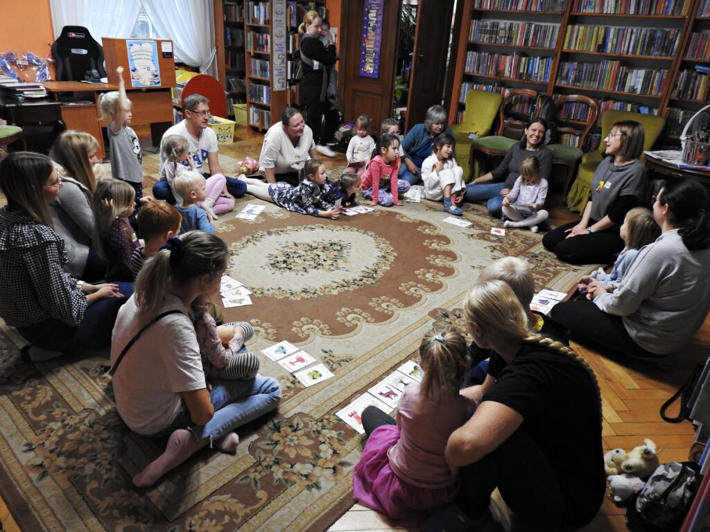 Duża grupa dzieci oraz dorosłych siedzi lub leży w kole na dywanie. Przed nimi leżą karty z różnymi grafikami. Wszyscy są skierowani w stronę bibliotekarki również siedzącej w kole. 1 dziecko unosi rękę.