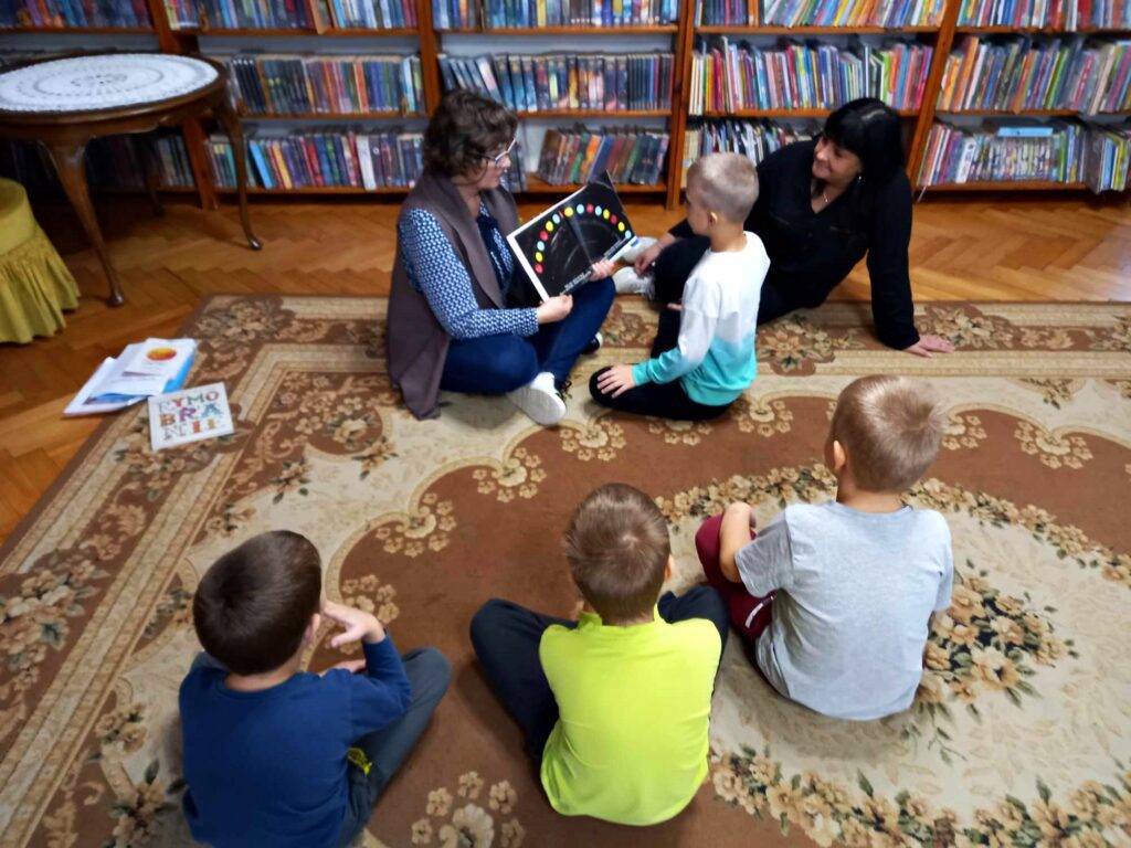 Dwie bibliotekarki siedzą na dywanie obok siebie. Jedna z nich trzyma otwartą książkę. Na stronach książki widać kolorowe kropki ułożone w półokręgu. Przed książką siedzi dziecko. Za nim 3 dzieci.