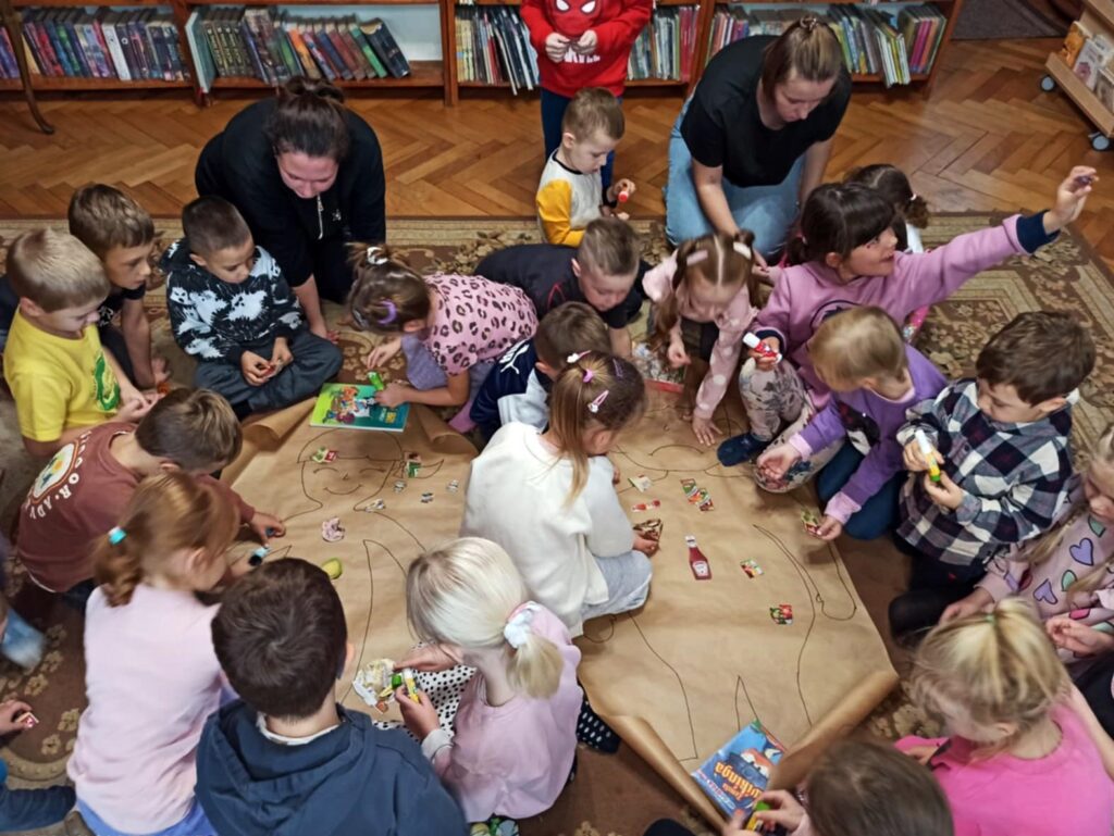 2 duże arkusze szarego papieru leżą na dywanie. Na jednym narysowana postać otyłego dziecka, na drugim szczupłego dziecka. Na postaciach naklejone są wycinki przedstawiające zdrowe i niezdrowe produkty spożywcze. Wokół arkuszy siedzi grupa kilkunastu dzieci oraz 2 osoby dorosłe. Dzieci naklejają wycinki lub przyglądają się.