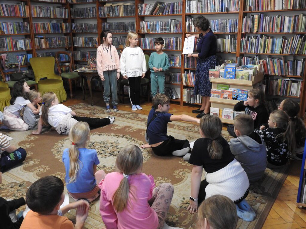 3 dzieci oraz bibliotekarka stoją. Przed nimi siedzi duża grupa dzieci. Bibliotekarka trzyma w ręku kartkę z nadrukowanymi grafikami. W tle regały z książkami.