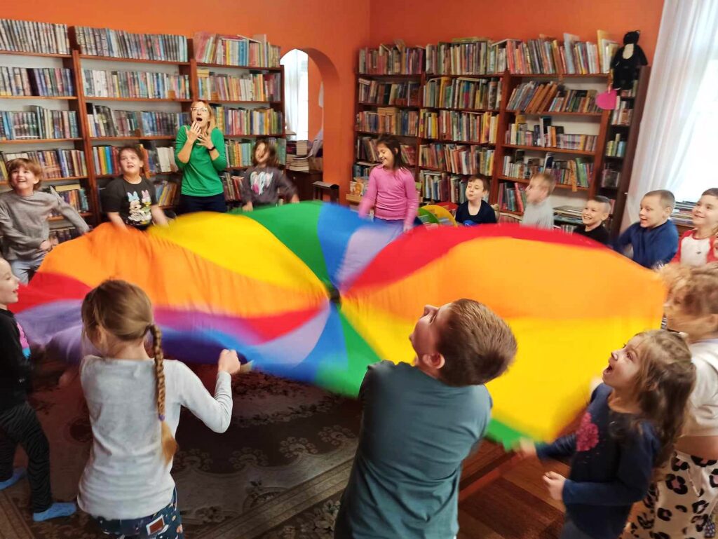 Grupa kilkunastu dzieci oraz bibliotekarka stoją wokół kolorowej chusty animacyjnej. Patrzą w górę. Dzieci śmieją się. Bibliotekarka trzyma dłonie przy twarzy, ma otwartą buzię w geście zdziwienia. W tle regały z książkami.