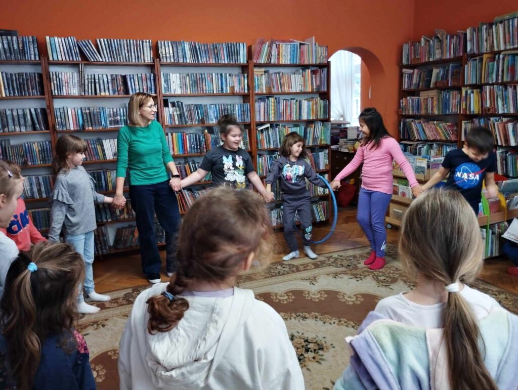 Grupa dzieci oraz bibliotekarka stoją w kole trzymając się za ręce. Pomiędzy dwójką dzieci, na złączonych rękach jest zawieszone niebieskie hula hop. W tle regały z książkami.