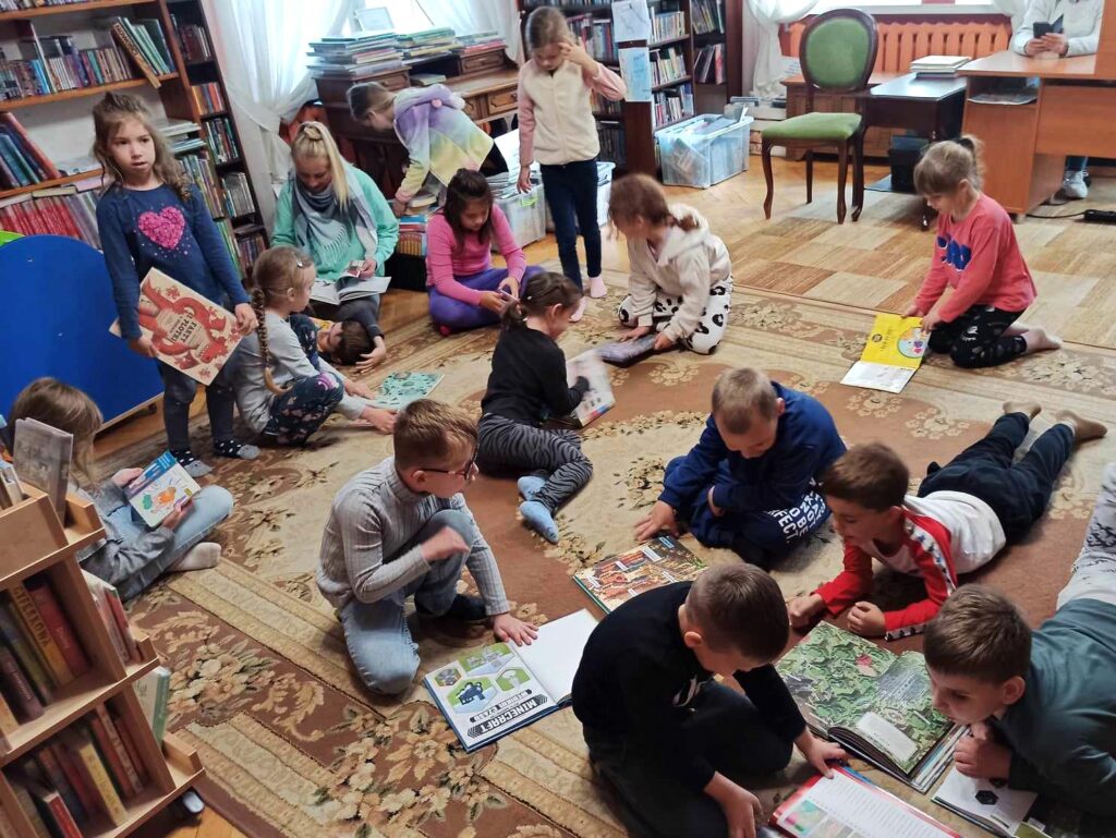 14 dzieci oraz 1 osoba dorosła są w bibliotece. Część z nich siedzi lub leży na dywanie, a część stoi. Oglądają książki.