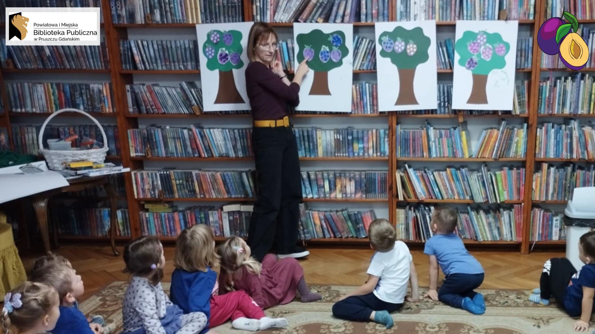 Na dywanie siedzą dzieci. dzieci i patrzą w stronę stojącej przed nimi bibliotekarki. Bibliotekarka pokazuje na przyklejoną na regale kartkę. Na białej kartce naklejone zdjęcie drzewa ze śliwkami.