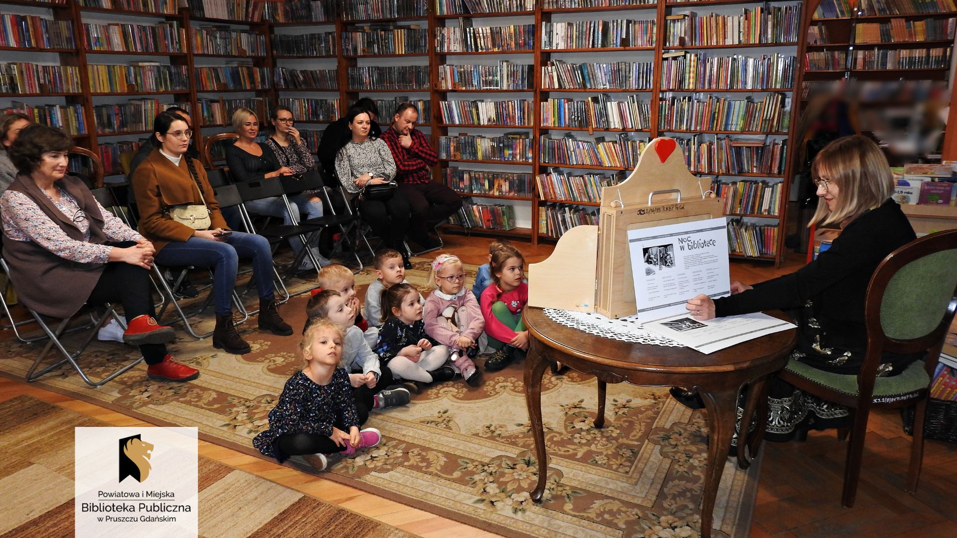 Grupa dzieci siedzi na dywanie. Za nimi na krzesłach siedzi 9 osób dorosłych. Przed nimi na krześle siedzi bibliotekarka. Obok bibliotekarki stoi okrągły stolik. Na stoliku znajduje się otwarta skrzynia teatrzyku kamishibai. Za skrzynią bibliotekarka trzyma kartę teatrzyku z tekstem i czarno-białą ilustracją. W tle regały z książkami.