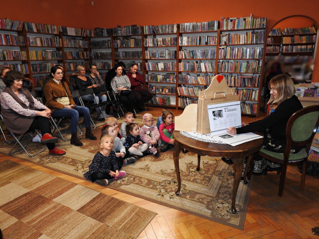 Grupa dzieci siedzi na dywanie. Za nimi na krzesłach siedzi 9 osób dorosłych. Przed nimi na krześle siedzi bibliotekarka. Obok bibliotekarki stoi okrągły stolik. Na stoliku znajduje się otwarta skrzynia teatrzyku kamishibai. Za skrzynią bibliotekarka trzyma kartę teatrzyku z tekstem i czarno-białą ilustracją. W tle regały z książkami.