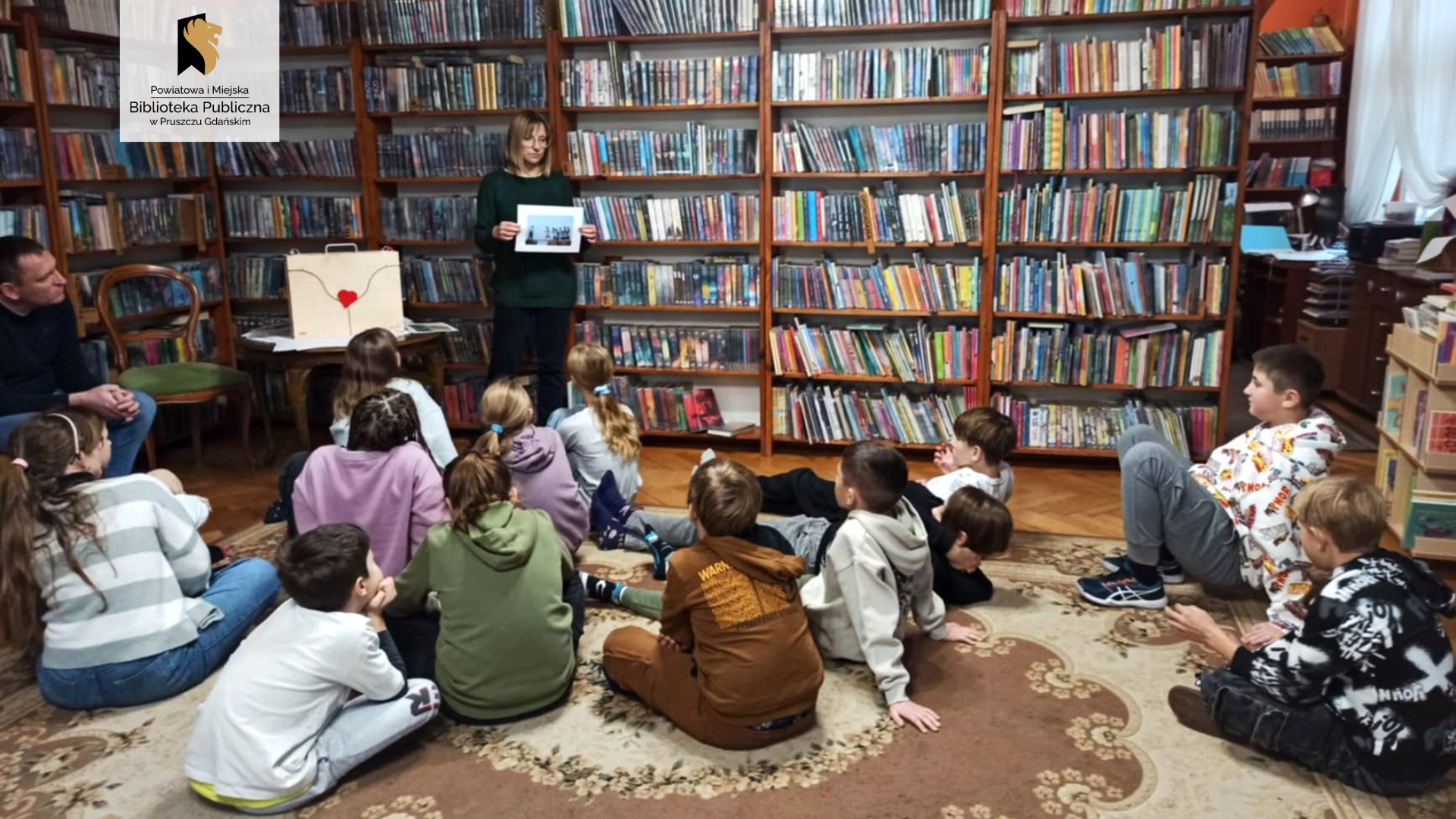 Grupa dzieci siedzi na dywanie. Są skierowani w stronę bibliotekarki, która stoi przed nimi i trzyma w ręku fotografię. Obok bibliotekarki jest stolik, na którym znajduje się zamknięta skrzynia teatrzyku kamishibai. W tle regały z książkami.