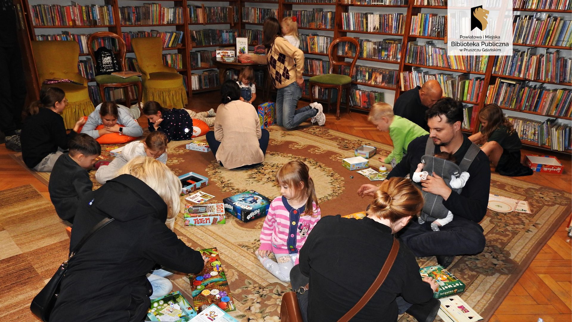 6 osób dorosłych i 11 dzieci jest w bibliotece. Większość siedzi lub leży na dywanie w grupach, wokół rozłożonych różnych gier planszowych. 2 osoby wybierają planszówki ze stolika.