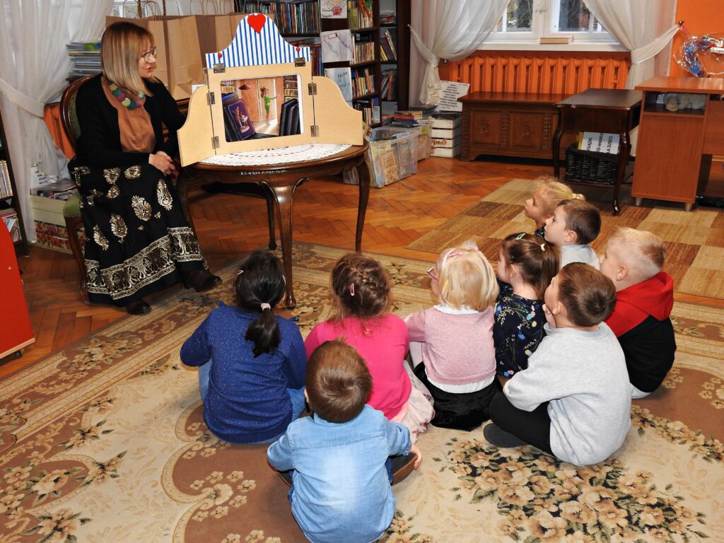 9 dzieci siedzi na dywanie. Są skierowane w stronę bibliotekarki siedzącej na krześle. Obok bibliotekarki znajduje się otwarta skrzynia teatrzyku kamishibai stojąca na stoliku. W skrzyni widoczna jest kolorowa ilustracja. 