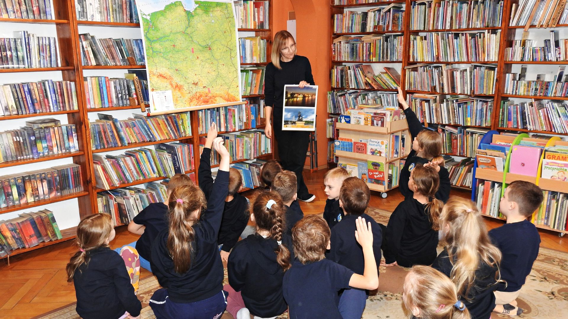 Bibliotekarka stoi obok fizycznej mapy Polski zawieszonej na regale z książkami. Trzyma w ręku kolaż dwóch zdjęć. Na jednym zdjęciu Warszawa nocą, na drugim Syrenka Warszawska. Przed bibliotekarką siedzi grupa dzieci. 4 dzieci unosi rękę.