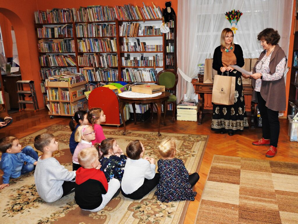 Dwie bibliotekarki stoją. Jedna trzyma w ręku dyplom. Druga bibliotekarka trzyma w dłoniach papierową torbę z logo biblioteki. Przed kobietami siedzi grupa 9 dzieci.